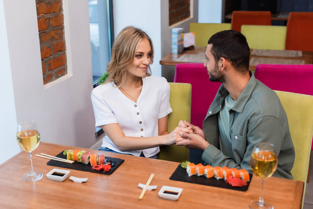 pareja feliz tomados de la mano y mirándose cerca de sushi delicioso en el restaurante - Foto, imagen
