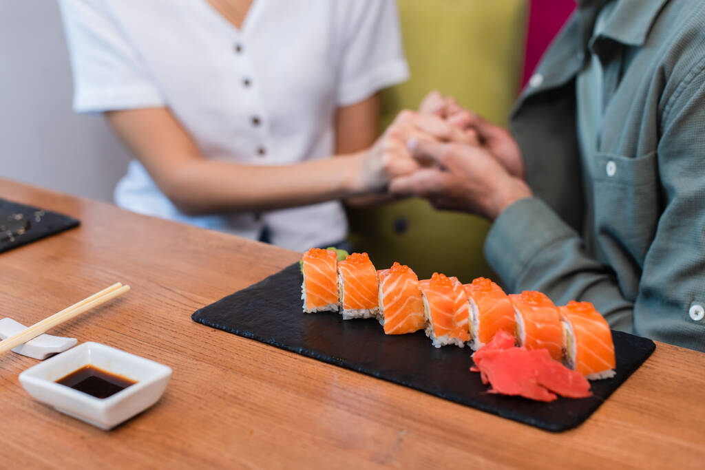 przycięty widok rozmytej pary trzymając się za ręce w pobliżu płyty z pysznymi bułkami sushi w restauracji - Zdjęcie, obraz