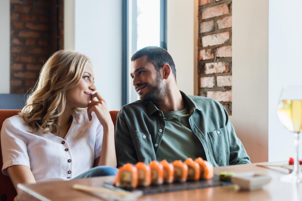 joyful couple smiling at each other near blurred sushi rolls and glass of white wine - Foto, Imagem