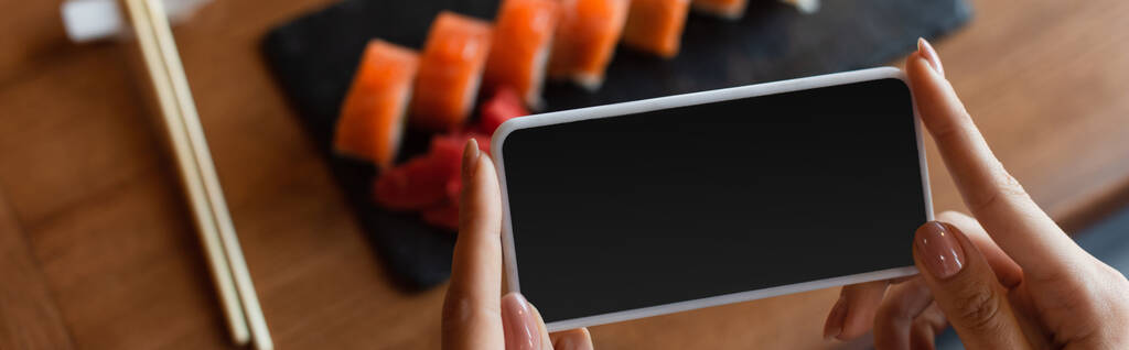 rajattu näkymä nainen tilalla älypuhelin tyhjä näyttö lähellä hämärtynyt sushi rullina, banneri - Valokuva, kuva