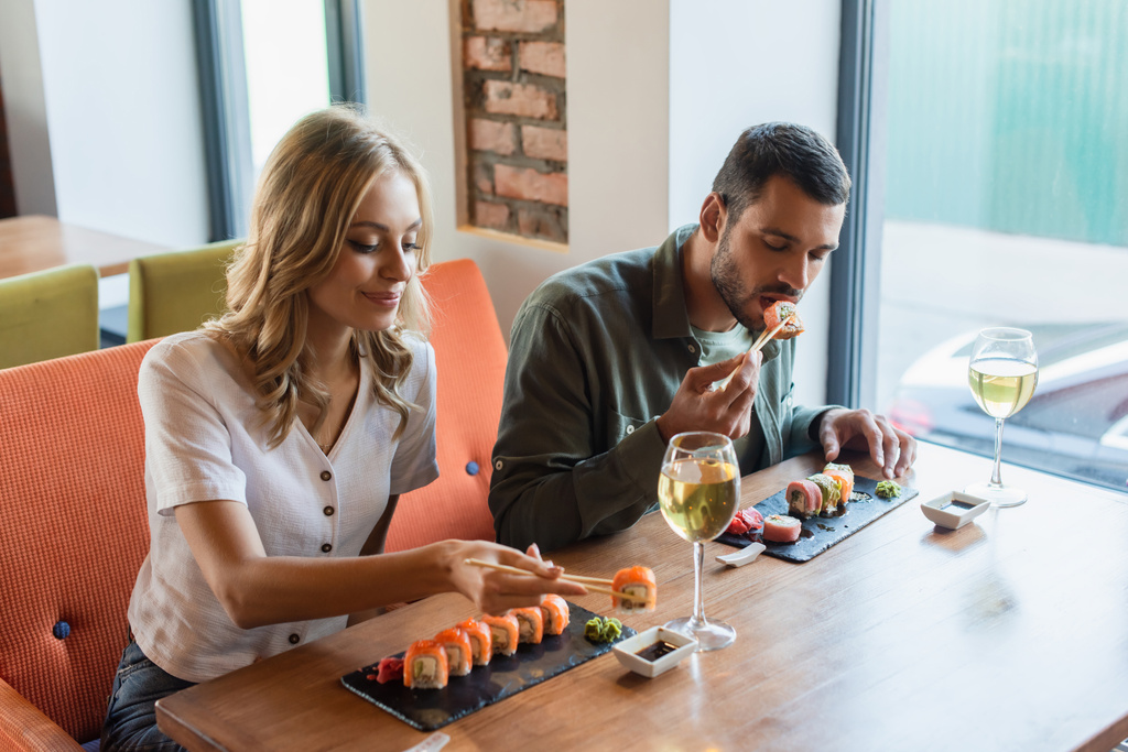 白ワインのグラスと彼氏の近くでおいしい寿司ロールを食べる幸せな女性 - 写真・画像