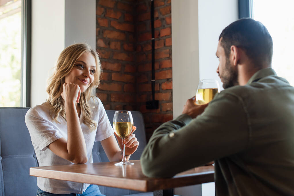 radosna kobieta patrząc na rozmazanego mężczyznę podczas picia białego wina w restauracji - Zdjęcie, obraz