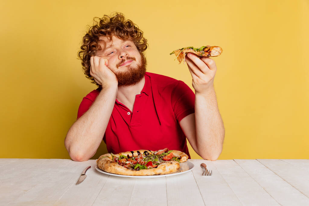 Portret van een roodbaarde man proeverij heerlijke Italiaanse pizza geïsoleerd op gele studio achtergrond. - Foto, afbeelding