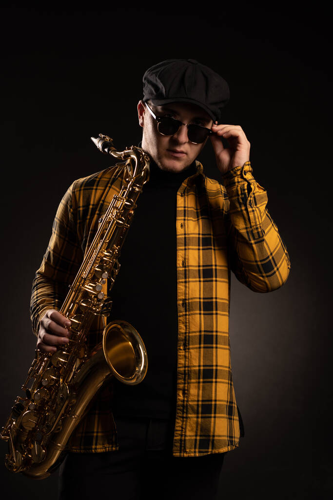 Hombre Saxofonista con Camisa Amarilla a Cuadros, Gorra y Elegantes Gafas de Sol sosteniendo un Alto Saxofón. Fondo negro. Primer plano Retrato - Foto, imagen