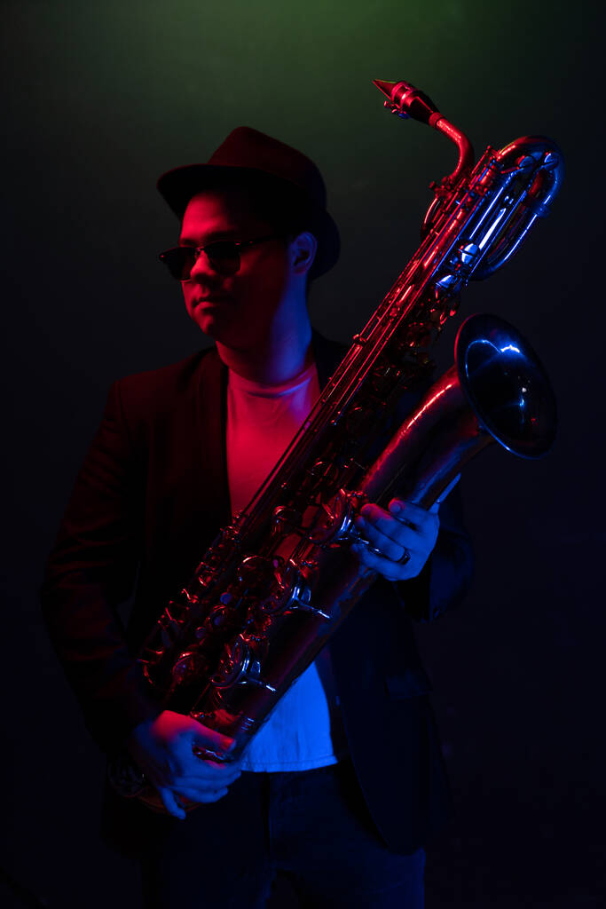 Young Saxophonist in a Black Jacket, Hat and Sunglasses Holds a Baritone Saxophone. Fondo oscuro. Luz de neón. Primer plano Retrato - Foto, imagen