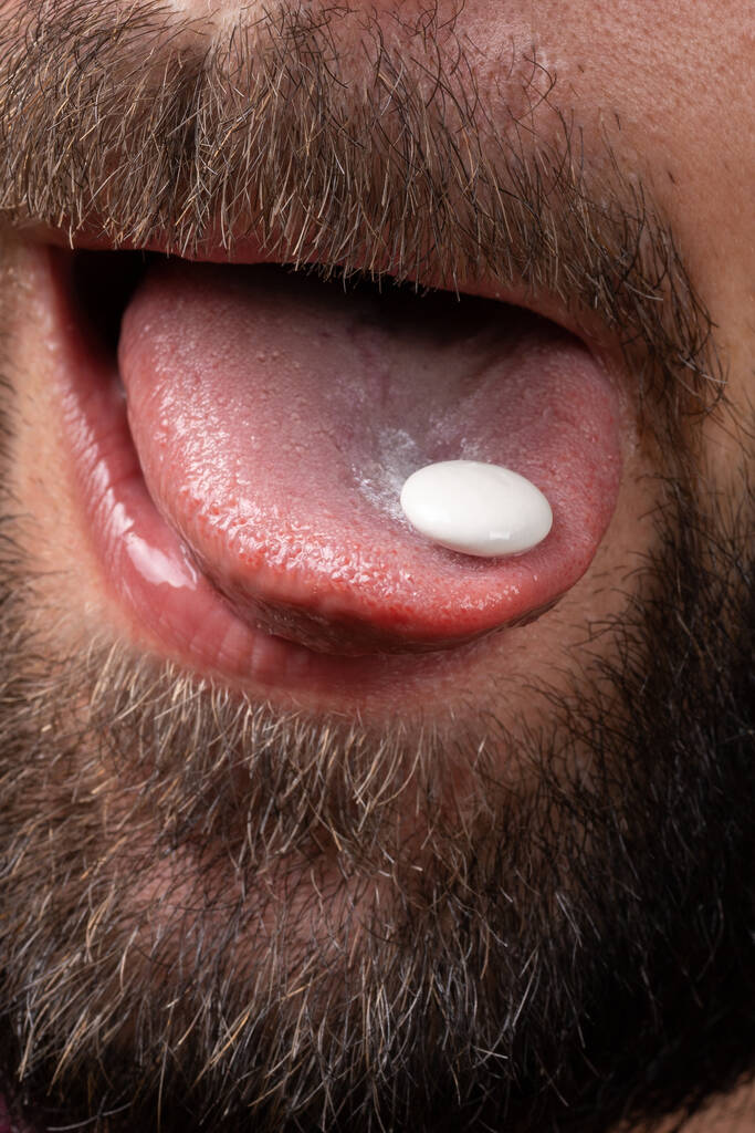 Екстремально крупним планом біла кругла таблетка на язиці бородатого чоловіка стикається з темними вусами, приймає вітаміни, знеболює
 - Фото, зображення