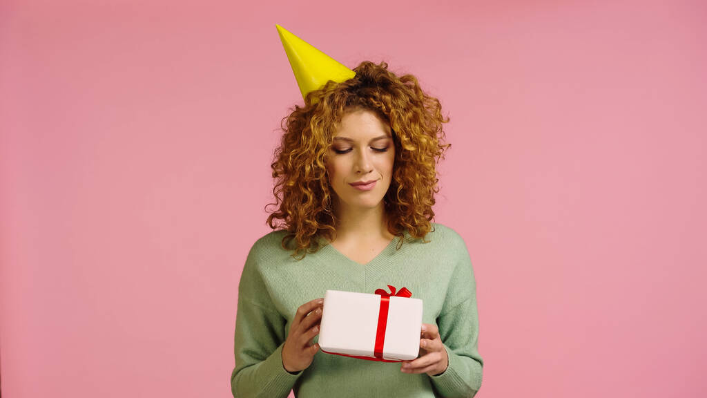 attente vrouw in party cap op zoek naar geschenk doos geïsoleerd op roze - Foto, afbeelding
