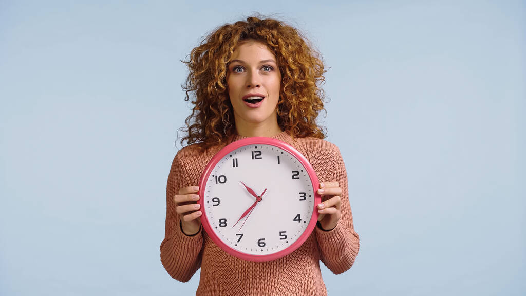 amazed redhead woman with open mouth holding round clock isolated on blue - Photo, Image