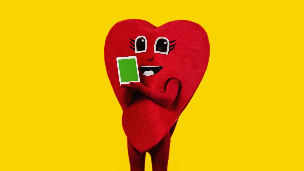 человек в красном костюме сердца держит цифровой планшет с зеленым экраном изолирован на желтый - Фото, изображение