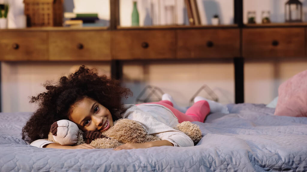 feliz afroamericano niño acostado con osito de peluche en la cama  - Foto, Imagen