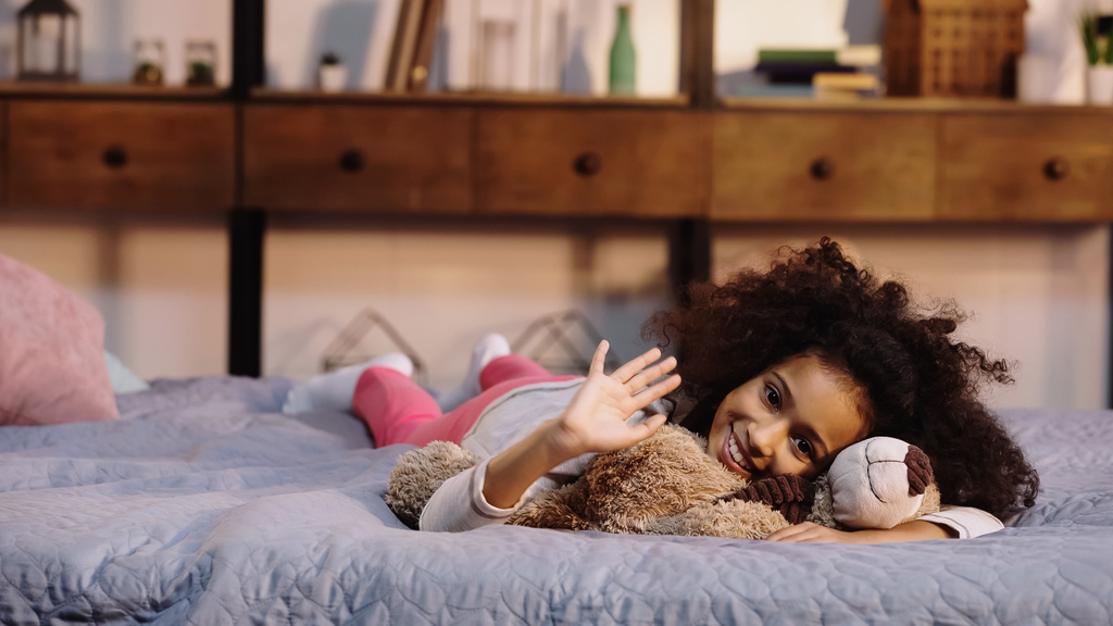 felice afro americano bambino agitando mano mentre sdraiato con orsacchiotto a letto
 - Foto, immagini