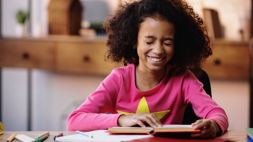 onnellinen afrikkalainen amerikkalainen tyttö lukee kirjaa ja nauraa kotona - Valokuva, kuva