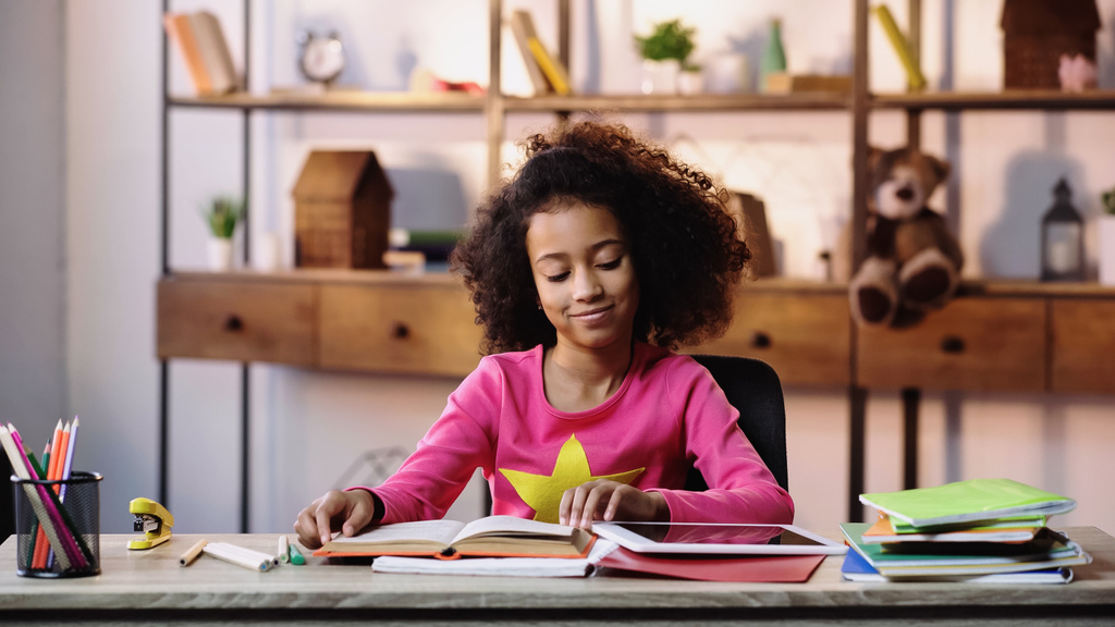 gelukkig Afrikaans amerikaans meisje lezen boek in de buurt van notebooks en digitale tablet op bureau - Foto, afbeelding