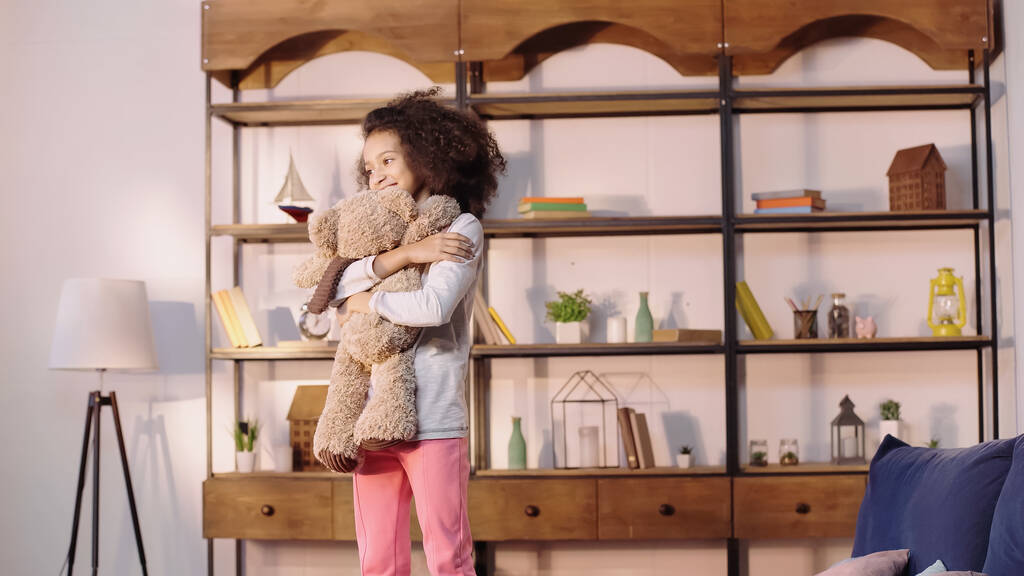 alegre africana americana chica abrazando teddy oso en sala de estar  - Foto, imagen