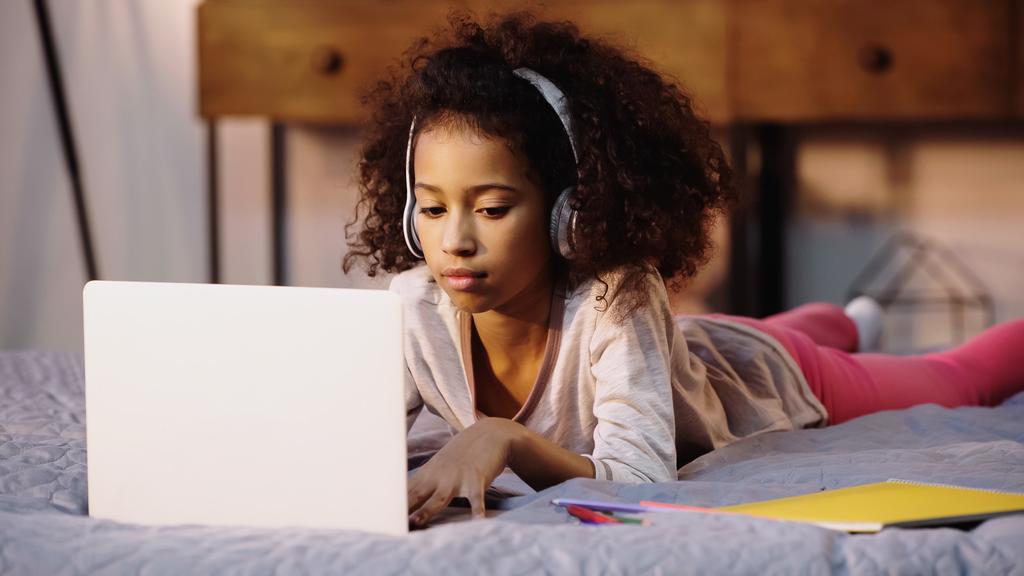 curly african american child in wireless headphones using laptop in bed - Фото, изображение