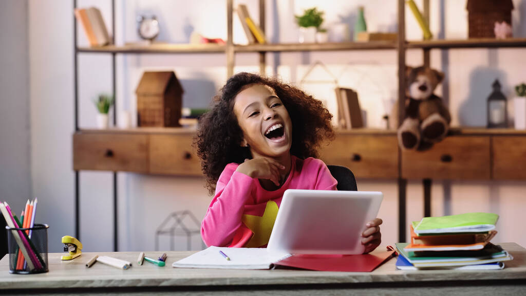 alegre afroamericana chica usando digital tablet y riendo cerca de cuadernos en escritorio  - Foto, imagen