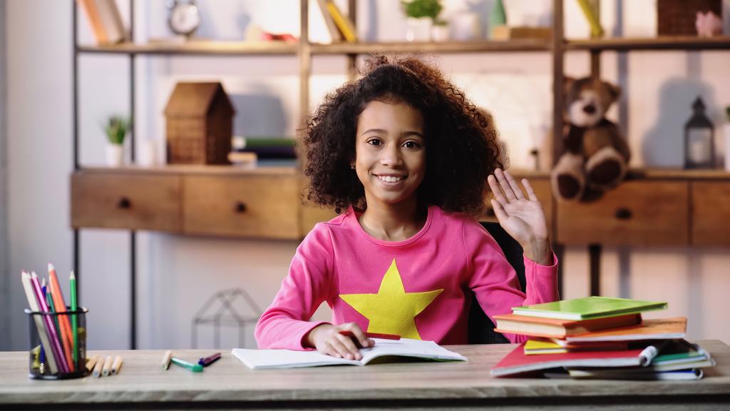 щаслива афроамериканська дівчина махає рукою і дивиться на камеру під час домашнього завдання
  - Фото, зображення