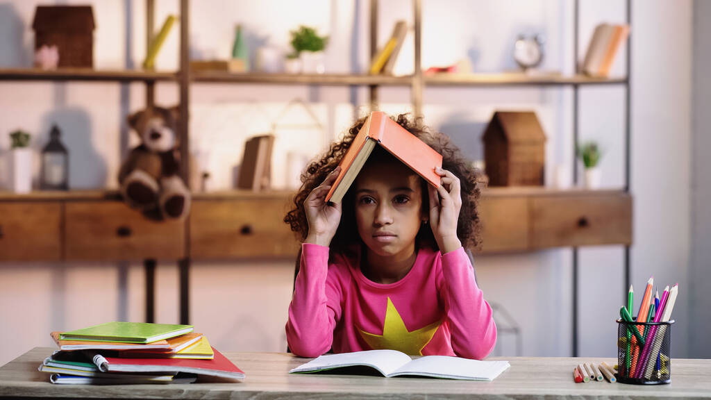 скучающий африканский американец держит книгу на голове, делая домашнее задание - Фото, изображение