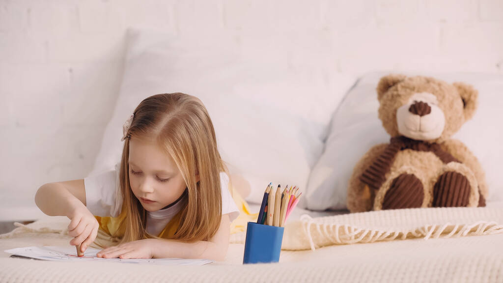 Детский рисунок с цветными карандашами рядом с размытым плюшевым мишкой на кровати  - Фото, изображение