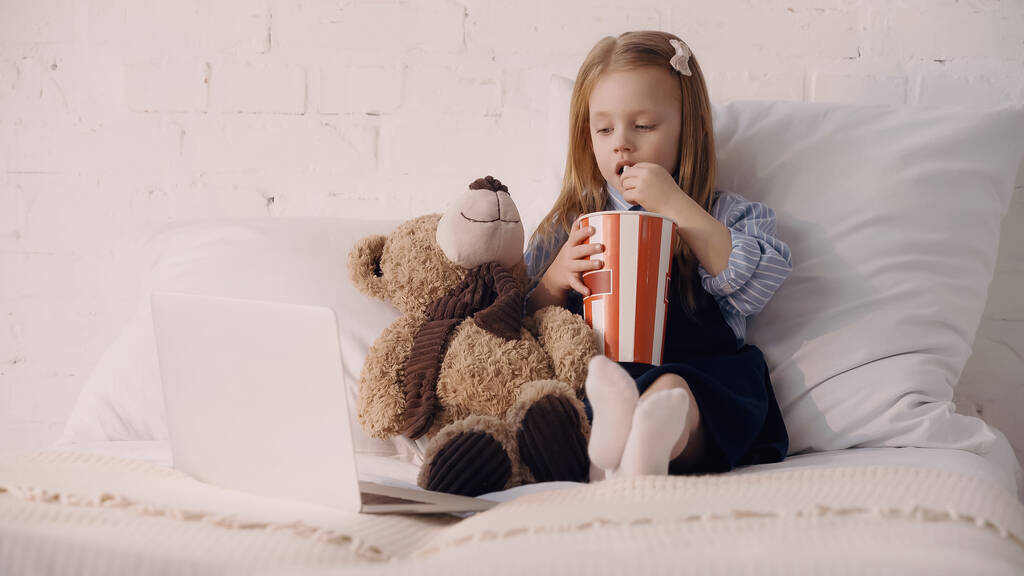 Kind isst Popcorn neben Teddybär und Laptop im Bett  - Foto, Bild