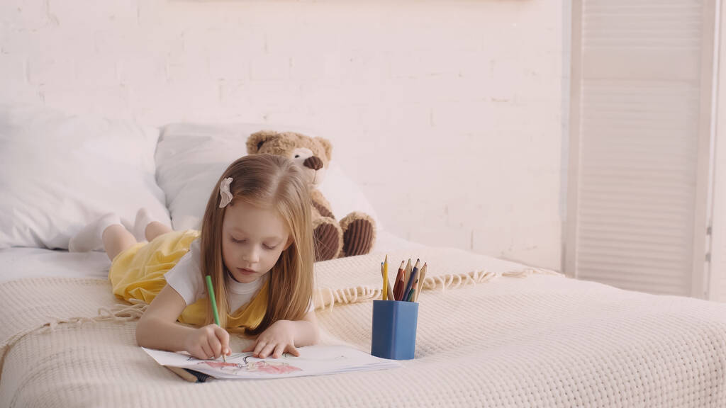 Kid tekening terwijl liggend op bed in de buurt van zacht speelgoed in de slaapkamer  - Foto, afbeelding