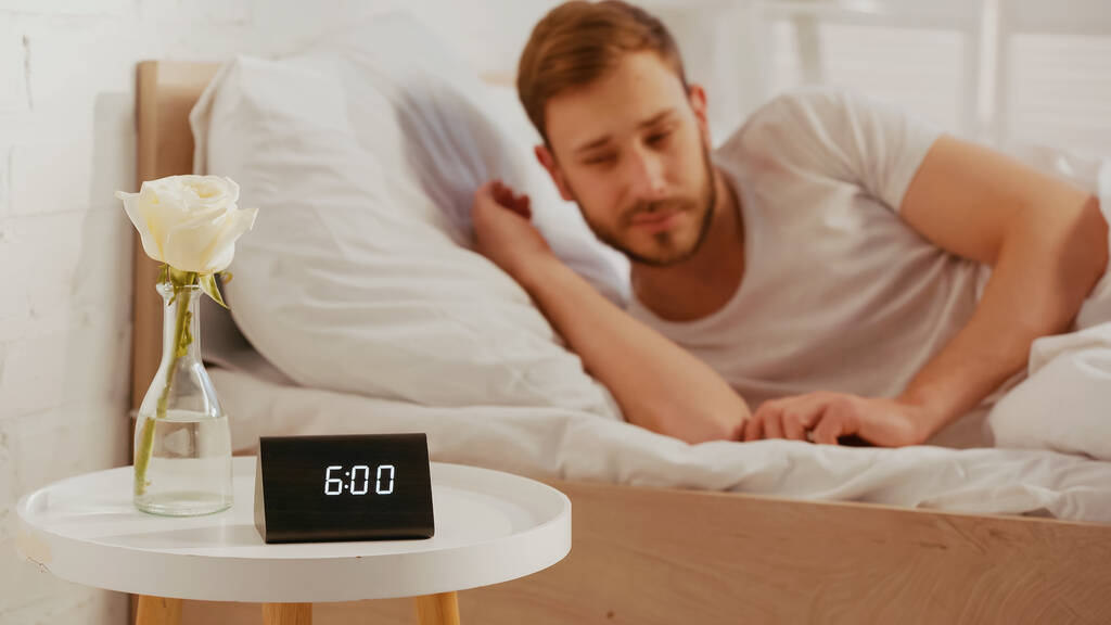 Годинник і квітка на тумбочці біля розмитого чоловіка на ліжку вдома
  - Фото, зображення