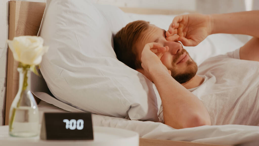 Hombre somnoliento frotando los ojos cerca de reloj borroso en la mesita de noche en casa  - Foto, imagen