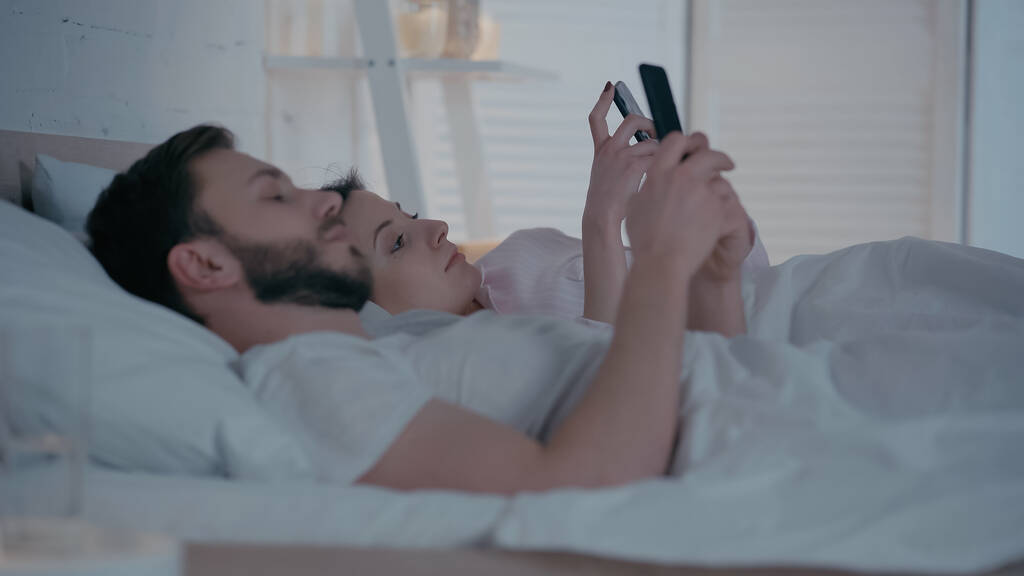 Νεαρή γυναίκα που χρησιμοποιεί smartphone κοντά στον σύζυγο στο κρεβάτι στο σπίτι  - Φωτογραφία, εικόνα