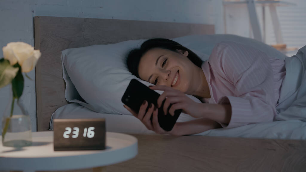 Cheerful woman using smartphone near blurred clock and plant in bedroom at night  - Photo, Image