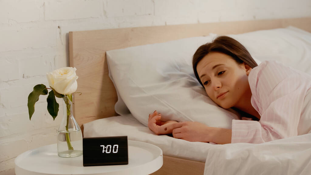 Mujer somnolienta mirando el reloj cerca de la flor en el dormitorio  - Foto, imagen