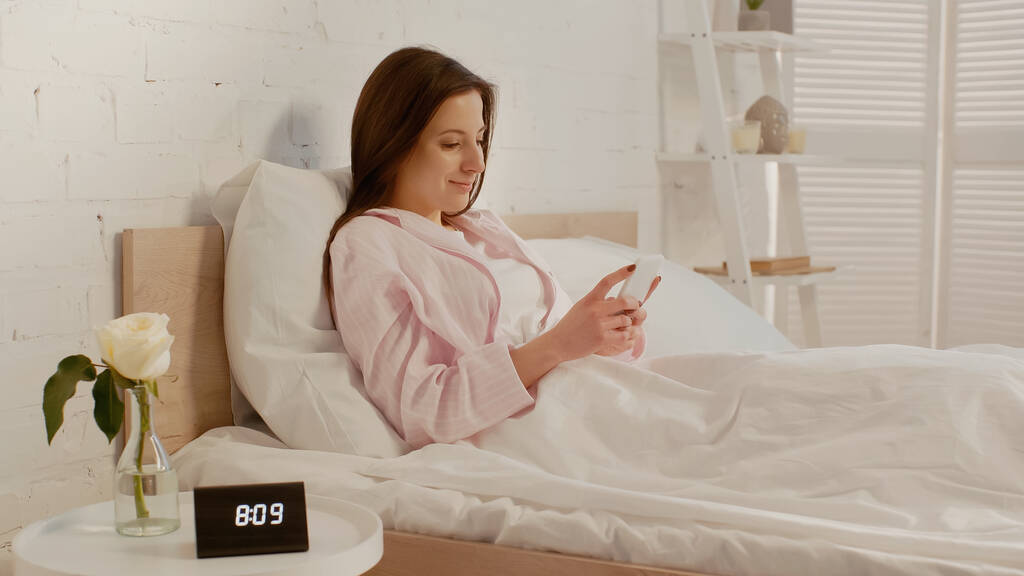 Glückliche Frau mit Smartphone auf Bett neben Uhr und Blume auf Nachttisch  - Foto, Bild