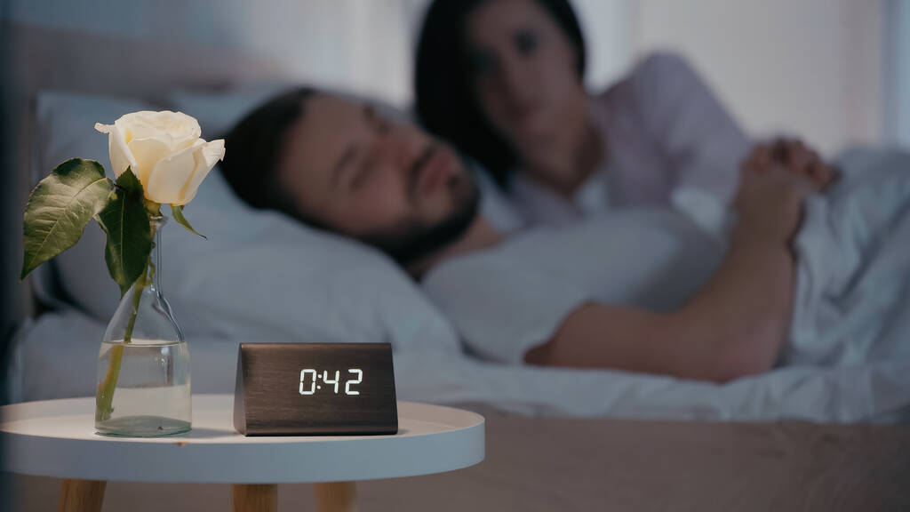 Planta y reloj cerca de pareja borrosa en la cama por la noche  - Foto, imagen