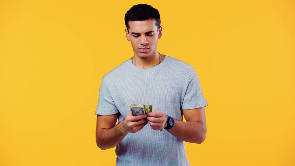 Tişörtlü genç adam sarıda izole edilmiş dolarları sayıyor.  - Fotoğraf, Görsel