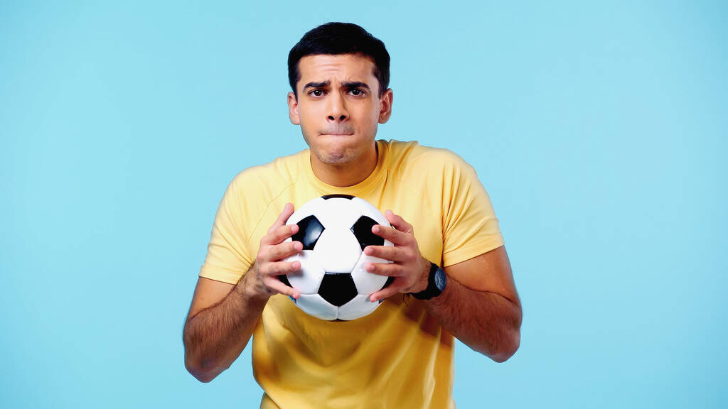 zmartwiony młody człowiek w żółtej koszulce trzyma piłkę nożną odizolowany na niebiesko - Zdjęcie, obraz
