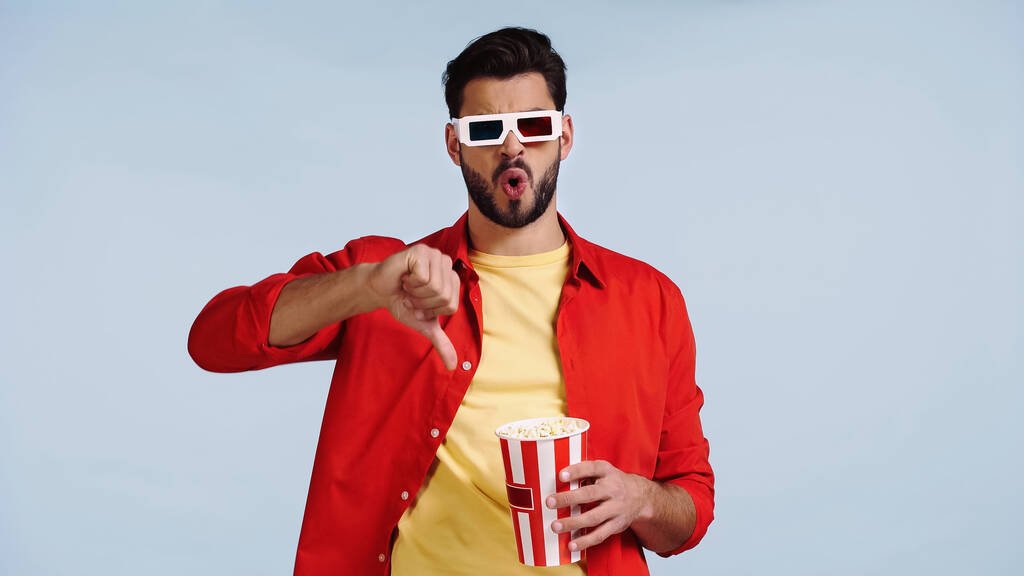 junger bärtiger Mann mit 3D-Brille schaut Film, zeigt Daumen nach unten und hält Popcorn-Eimer isoliert auf blauem Grund  - Foto, Bild