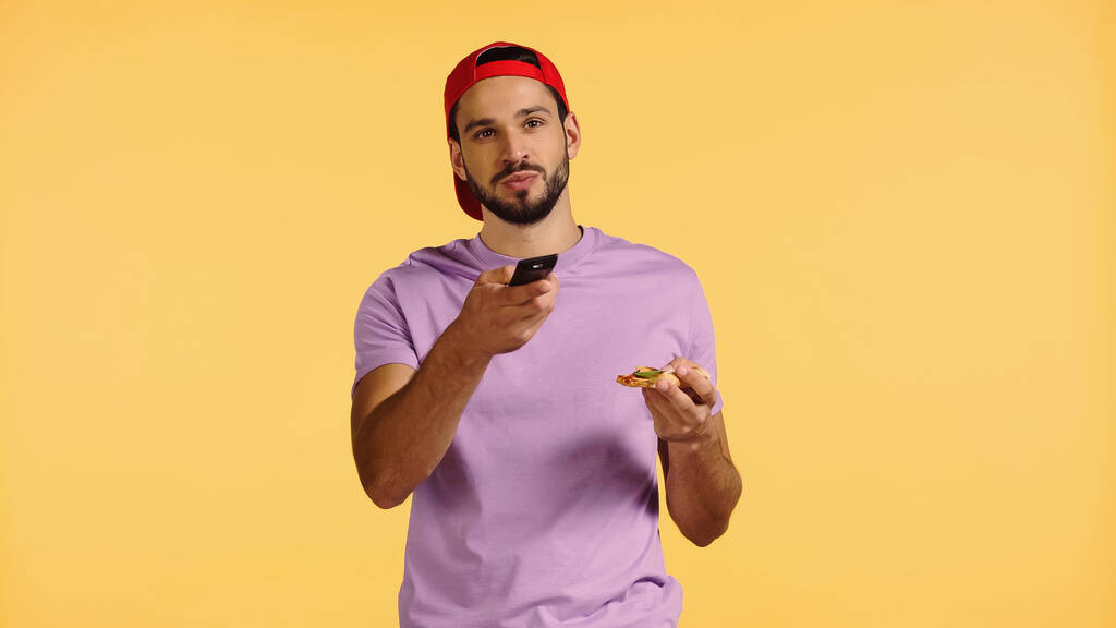 γενειοφόρος άνδρας που κρατάει πίτσα και ενεργοποιεί κανάλια απομονωμένα στο κίτρινο  - Φωτογραφία, εικόνα