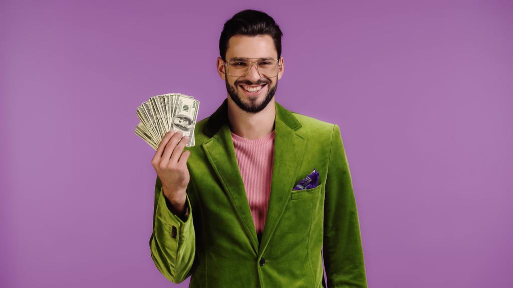 happy man in green jacket and glasses holding dollars isolated on purple - Fotó, kép