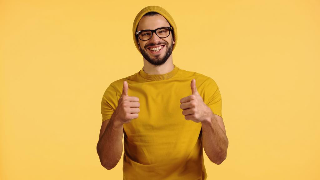 happy young man in beanie hat, glasses and t-shirt showing thumbs up isolated on yellow - Фото, изображение