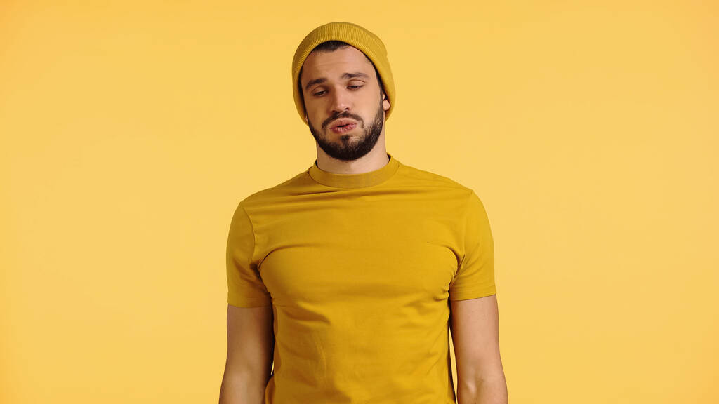 tired young man in beanie hat and t-shirt puffing cheeks isolated on yellow - Foto, Bild