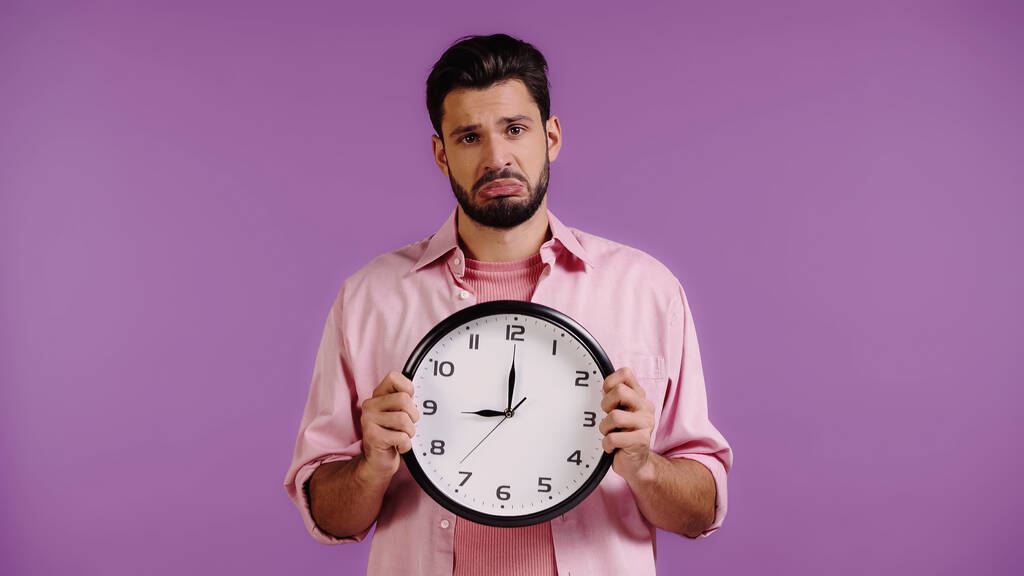 bouleversé jeune homme tenant horloge isolé sur violet  - Photo, image