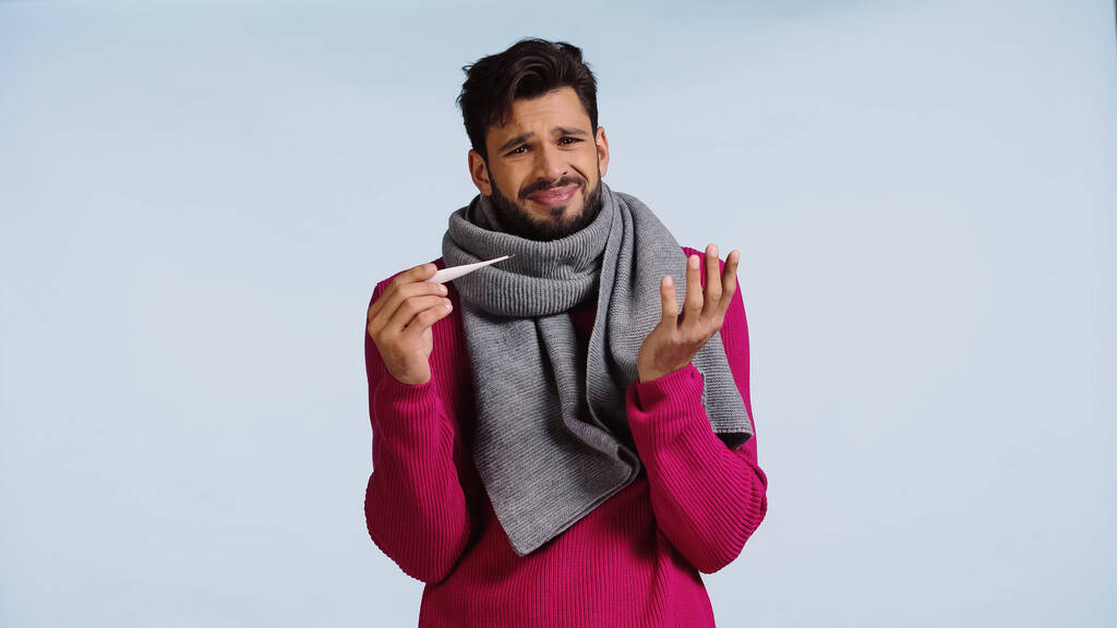 diseased man in pink sweater and scarf holding digital thermometer isolated on blue  - Фото, изображение
