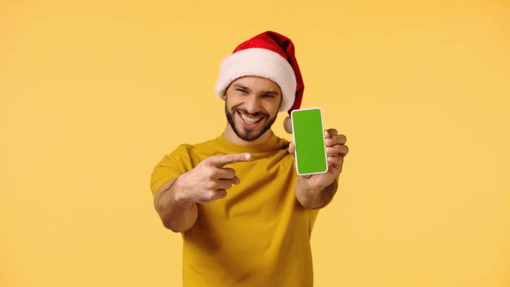 счастливый человек в санта-шляпе, указывающий на смартфон с зеленым экраном, изолированным на желтом - Фото, изображение