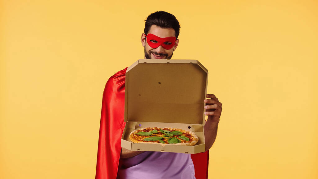 happy man in superhero costume opening box with pizza isolated on yellow - Фото, изображение