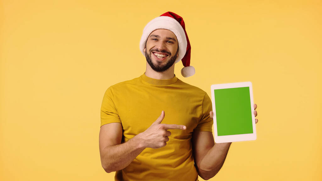 homem feliz em santa chapéu apontando com o dedo para tablet digital isolado em amarelo - Foto, Imagem