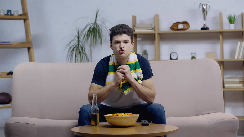 worried sport fan in striped scarf watching championship on sofa near beer and chips - Foto, immagini