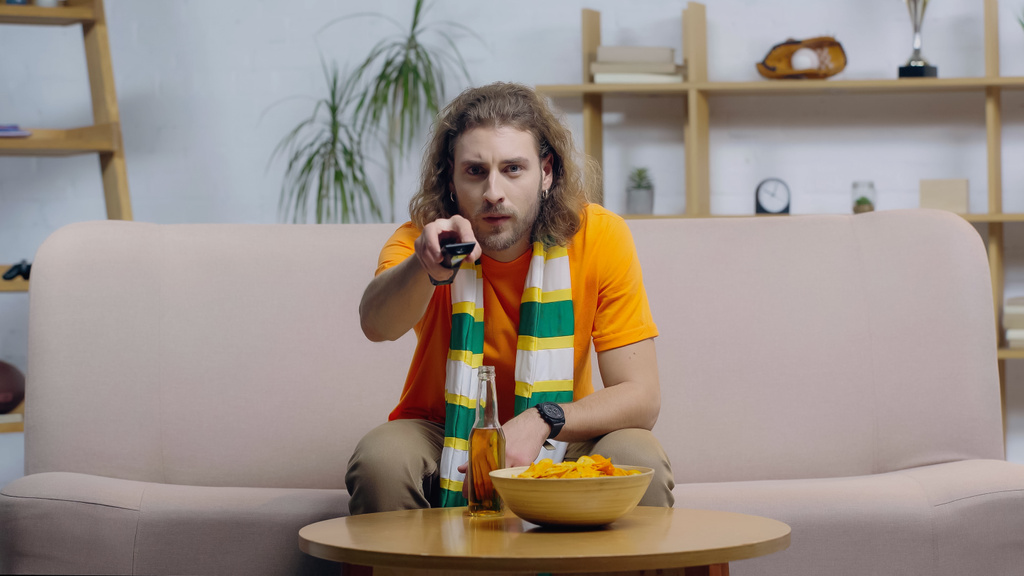 вентилятор в полосатом шарфе, щёлкающий по телеканалам, сидя рядом с пивом и чипсами на кофейном столике - Фото, изображение
