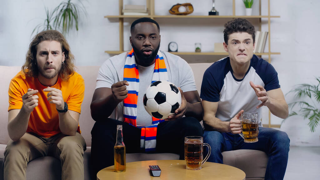 african american man with soccer ball holding clenched fist for luck while watching football match with worried friends - Foto, Imagem