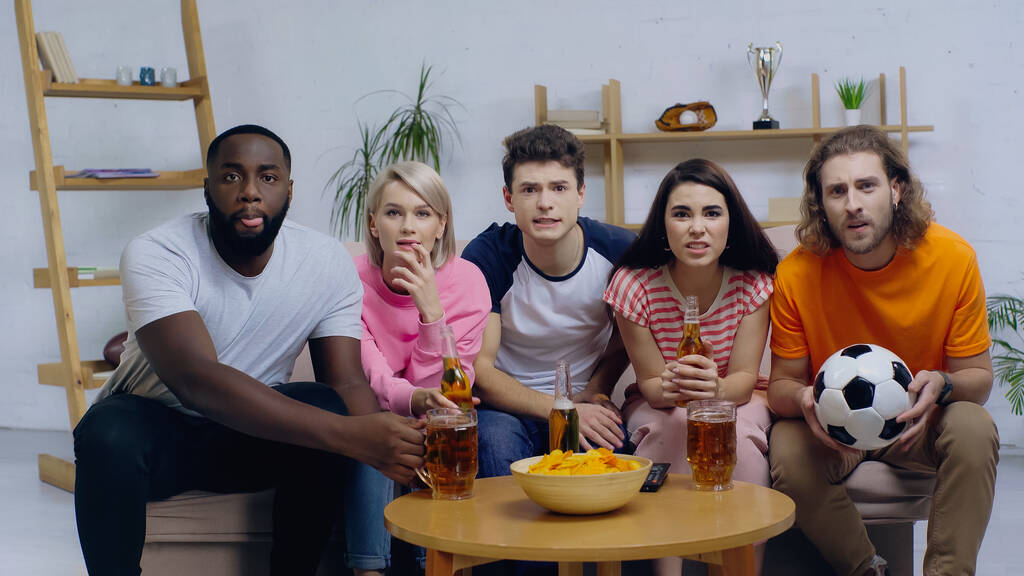 tense multiethnic friends sitting on sofa near beer and chips while watching football match - Photo, Image
