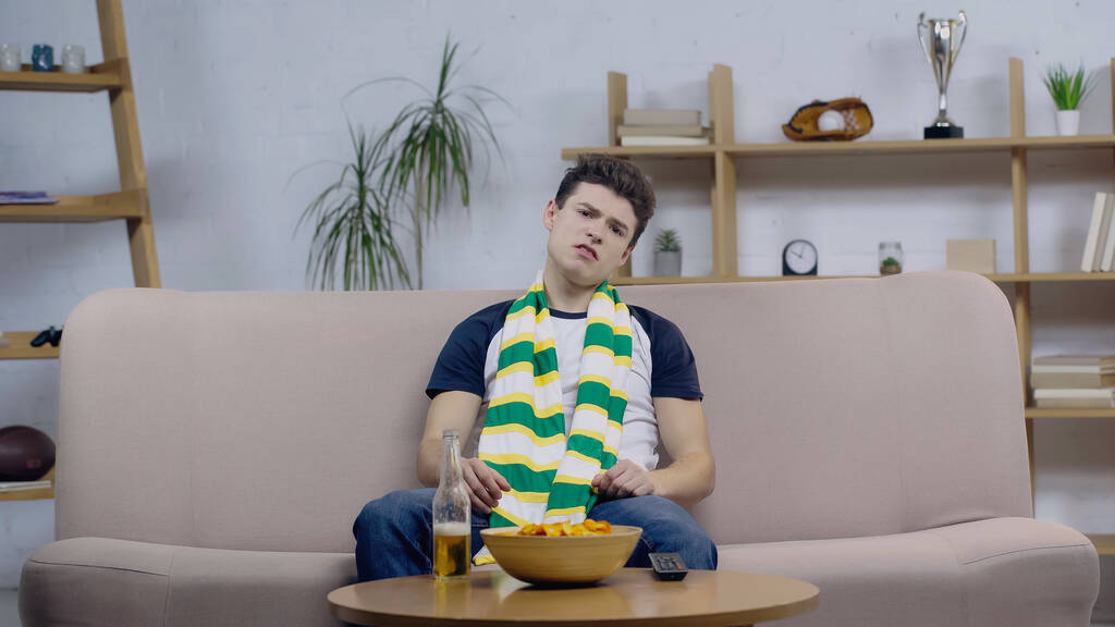 frustrated sport fan sitting on couch near beer and chips while watching game on tv - Foto, immagini