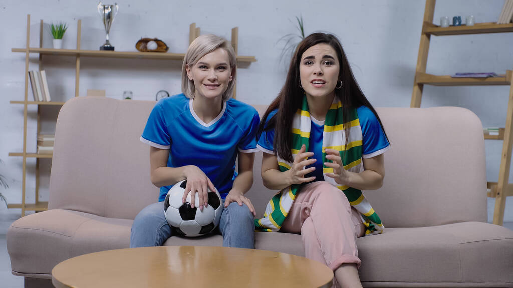 vzrušené ženy sledující fotbalové mistrovství, zatímco sedí na pohovce s fotbalovým míčem - Fotografie, Obrázek